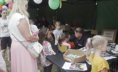 Na zdjęciu widać grupę dzieci i dorosłych przy stoisku z malowaniem twarzy. Kobieta maluje twarz jednej z dziewczynek, a inne dzieci i dorośli stoją obok i czekają. Nad stoiskiem wiszą kolorowe balony.