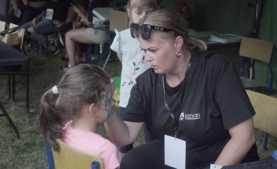 Na zdjęciu widzimy kobietę, która maluje twarz małej dziewczynce siedzącej na krześle. Kobieta jest skupiona na swojej pracy, ma blond włosy związane w kucyk, nosi czarną koszulkę z logo „Strzelecki Ośrodek Kultury” oraz okulary przeciwsłoneczne na głowie. Dziewczynka, której maluje twarz, ma ciemne włosy związane w kucyk i różową koszulkę. W tle stoi druga dziewczynka obserwująca tę scenę. Za nimi widać innych ludzi siedzących i zajętych różnymi aktywnościami.
