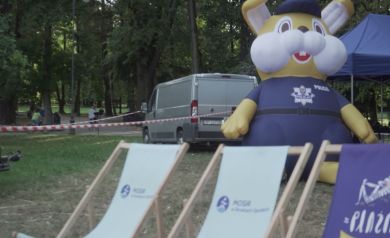 Na zdjęciu widzimy park z kilkoma leżakami z napisem „MOSiR w Strzelcach Opolskich” na pierwszym planie. Obok stoi duża, nadmuchiwana postać maskotki policji w granatowym mundurze i czapce. W tle jest szary samochód dostawczy oraz niebieski namiot. 