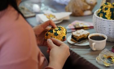 Kobieta w dłoni trzyma jajko ozdobione kolorowymi dekoracjami kwiecistymi. Przed nią na stole kołacz i kawa oraz koszyk już pełen tego rodzaju jajek. 