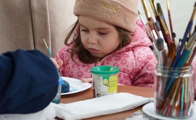 Mała dziewczynka w różowej kurtce i beżowej czapce , maluje gipsową ozdobę wielkanocną. 