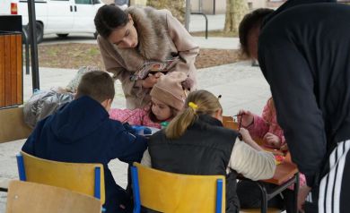 Dzieci przy stole wspólnie z rodzicami ozdabiają gipsowe jajeczka podczas warsztatów plastycznych na jarmarku.