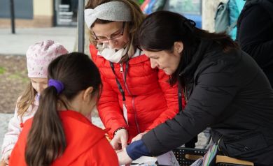 Panie prowadzące warsztaty z jajem, pomagają dziewczynce w czerwonej kurtce ozdabiać wielkanocne kartki.