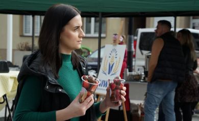 Kobieta z brązowymi włosami i  zielonym golfie idzie z kubeczkami pełnymi truskawek w czekoladzie, w tle food truck.