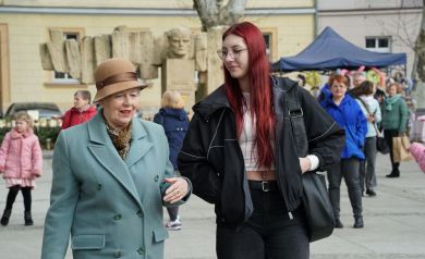 Starsza Pani  w zielonym płaszczu idzie z młodą dziewczyną w czerwonych włosach, w tle stoiska jarmarku. 