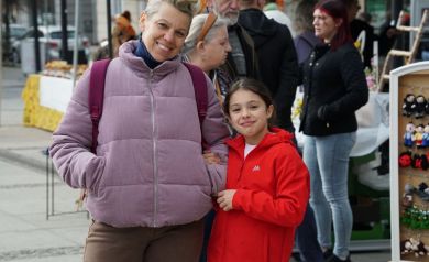 Mama w fioletowej kurtce z córeczką w czerwonej kurtce , uśmiechnięte patrzą w obiektyw. W tle stoisko z miodami.