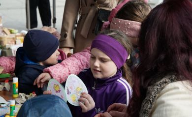 Dziewczynka w fioletowej opasce i bluzce ogląda efekty swojej pracy, kolorowa kartka wielkanocna w kształcie jajka. W tle inne dzieci podczas warsztatów. 
