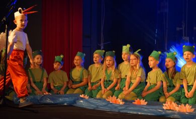 chłopiec przebrany za bociany biega na środku sceny, za nim siedzą przedszkolaki przebrane za żabki