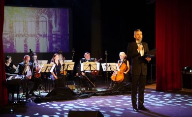 Na środku sceny mężczyzna rozpoczynający Galę Noworoczną. W tle orkiestra. 