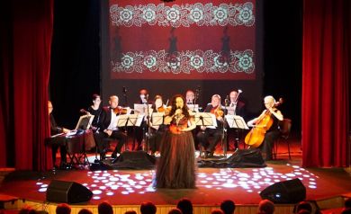 Na środku sceny śpiewaczka, w tle orkiestra grająca na różnych instrumentach. W tle dominuje czerwone światło sceniczne. 