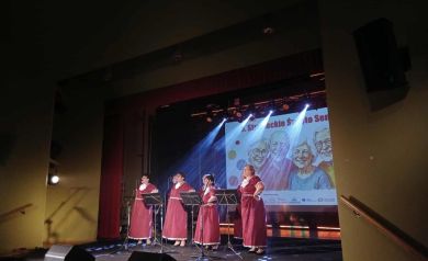 Na scenie występ zespołu śpiewaczego. Kobiety ubrane są w długie czerwone suknie z białymi dodatkami.