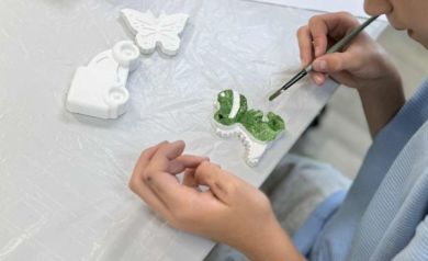 Chłopiec z pędzlem w ręku maluje zieloną farbą figurkę dinozaura. Obok niego na stole leżą również figurka przedstawiająca samochód i motyla. Stół jest pokryty folią. 