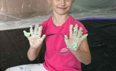 Dziewczynka w różowej bluzce siedzi na podłodze pokrytej czarną folią. Pokazuje do zdjęcia ubrudzone na zielono ręce. Przed nią pojemniki z różnymi produktami. W tle widać pomieszczenie pokryte folią. 