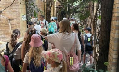 Dzieci biorące udział w wycieczce wśród zielonych roślin i pnączy. Widać tam również ceglane ściany.