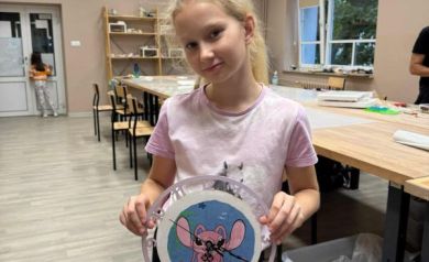 Na zdjęciu dziewczynka ubrana w różową koszulkę prezentuje wykonany przez siebie zegar. W tle widać innych uczestników warsztatów i sale, gdzie obywały się zajęcia. 