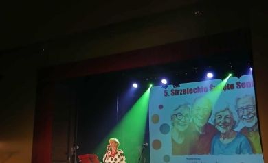 Na scenie kobieta ubrana w białą sukienkę w czarne duże kropki. Śpiewa do mikrofonu.