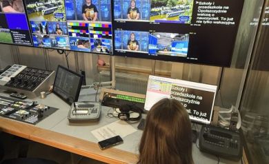 Na zdjęciu pomieszczenie z wieloma ekranami znajdujące się w budynku TVP 3 Opole. Przy komputerze siedzi młoda dziewczyna w fioletowej koszulce projektowej.  - powiększ