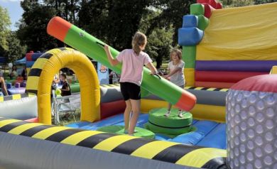 Na zdjęciu widać dwie dziewczynki bawiące się na dmuchańcu, stojąc na ruchomych platformach i walcząc piankowymi pałkami. 