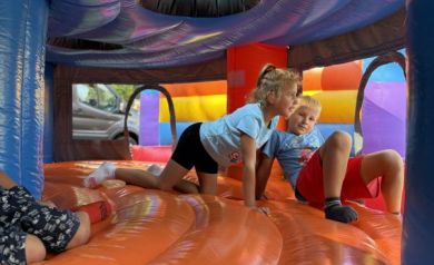 Na zdjęciu widzimy dwoje dzieci bawiących się wewnątrz dmuchanego zamku. Dzieci są na kolanach na pomarańczowej, nadmuchanej powierzchni. Dziewczynka ubrana jest w jasnoniebieską koszulkę i czarne spodenki, a chłopiec ma na sobie jasnoniebieską koszulkę i czerwone spodenki. W tle widać kolorowe ściany dmuchanego zamku oraz zewnętrzne otwory, przez które widać samochód i zieleń na zewnątrz.