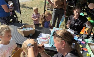 Na zdjęciu widać kobietę malującą twarz małej dziewczynce siedzącej na krześle. Wokół stoi kilka innych dzieci, które czekają na swoją kolej. Cała scena dzieje się pod namiotem udekorowanym kolorowymi balonami. W tle widoczne są stoły z farbami i innymi materiałami do malowania twarzy. 