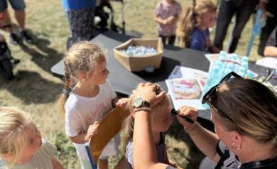 Na zdjęciu widać stanowisko malowania twarzy podczas wydarzenia „Zakręcona Wakacyjna Meta”. Kobieta maluje twarz dziewczynce siedzącej na krześle, a obok czekają kolejne dzieci. W tle widoczne są balony, stolik z różnymi materiałami (cukierki, woda) i rodzice. 