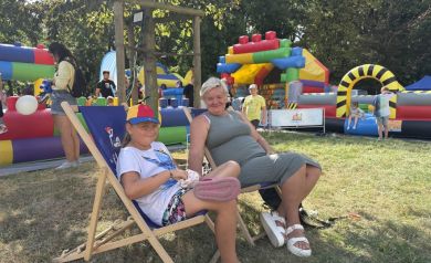 Na zdjęciu widać dwie osoby – dziewczynkę i kobietę, które siedzą wygodnie na leżakach na trawie. Dziewczynka ma na sobie kolorową czapkę z daszkiem, białą koszulkę i krótkie spodenki, natomiast kobieta ubrana jest w szarą sukienkę i białe sandały. W tle znajduje się kolorowe dmuchańce i inne atrakcje dla dzieci. Wokół jest jasno i słonecznie. W tle widać też inne dzieci i dorosłych.