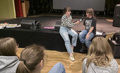 Na zdjęciu kobieta przeprowadzająca wywiad z uczestniczką projektu. Na pierwszym planie widać również resztę osób biorących udział w spotkaniu - są odwrócone tyłem do aparatu.  - powiększ