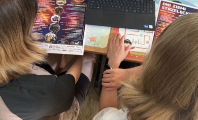 Na zdjęciu dwie młode dziewczyny pracujące na laptopie. Przed nimi plakaty z Dni Ziemi Strzeleckiej.  - powiększ
