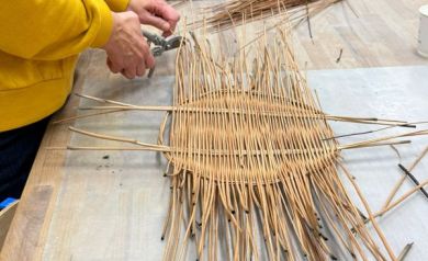 Na zdjęciu widać uczestników warsztatów wiklinarskich, którzy z zaangażowaniem wyplatają tacki katalońskie z cienkich gałązek wikliny przy dużym stole roboczym. 