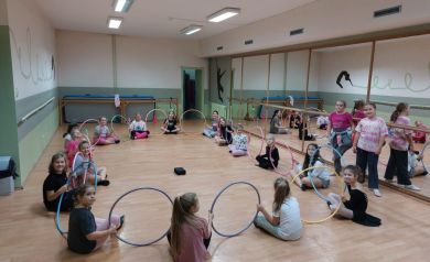 Na zdjęciu widać grupę uśmiechniętych dzieci podczas zajęć tanecznych w sali z lustrami, siedzących w kręgu z kolorowymi hula-hop. Atmosfera jest radosna i pełna energii.