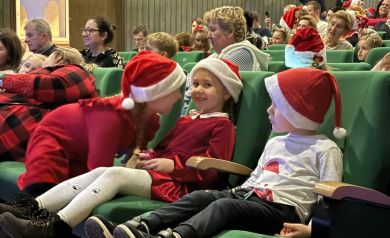 Na pierwszym planie trójka dzieci, dziewczynka nachyla się do drugiej dziewczynki. W tle publiczność. 