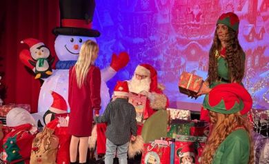 Na scenie są dzieci podchodzące po prezenty do mężczyzny przebranego za Mikołaja. Obok znajdują się dwie kobiety przebrane za elfy. Na zdjęciu widać również dekoracje świąteczne oraz liczne prezenty. 