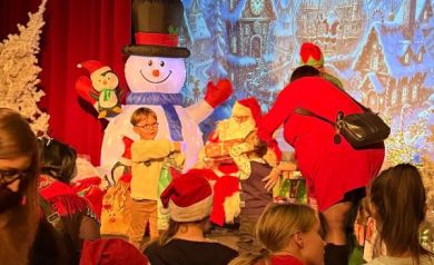 Dzieci podchodzące po  prezenty do mężczyzny przebranego za Mikołaja. W tle dekoracje świąteczne. 