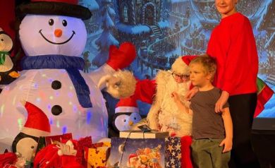 Młoda kobieta i chłopiec pozują do zdjęcia z mężczyzną przebranym za Mikołaja. Wokół nich znajdują się prezenty i świąteczna sceneria. 