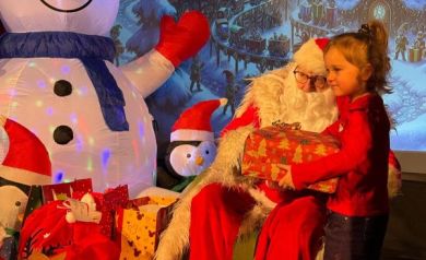 Dziewczynka pozująca do zdjęcia z mężczyzną przebranym za Mikołaja. Dziecko trzyma w dłoniach prezent. W tle widać świąteczną scenerię. 