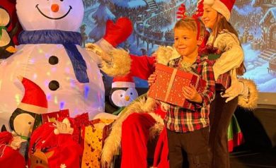 Chłopiec z prezentem . Za chłopcem widać dziewczynkę również z prezentem. W tle mężczyzna przebrany za Mikołaja i dekoracje świąteczne. 