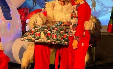 Chłopiec pozujący do zdjęcia z mężczyzną przebranym za Mikołaja. W tle świąteczna sceneria. 