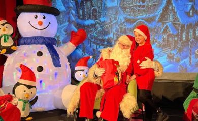 Dziecko i mężczyzna pozujący do zdjęcia z mężczyzną przebranym za Mikołaja. W tle widać świąteczną scenerię. 