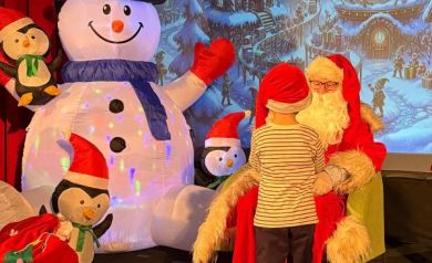 Dziecko odbierające prezent od mężczyzny przebranego za Mikołaja, siedzącego na zielonym fotelu. Na zdjęciu widać również dekoracje świąteczne. 