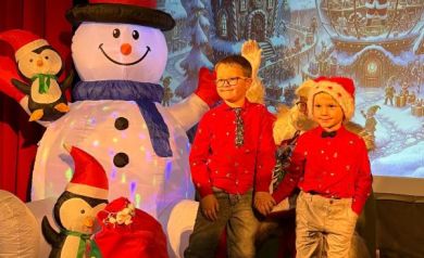 Na pierwszym pplanie widać dwóch chłopców trzymających się za dłonie. Za nimi mężczyzna przebrany za Mikołaja. W tle znajduje się dmuchana dekoracja bałwana i pingwiny oraz duży worek z prezentami. 