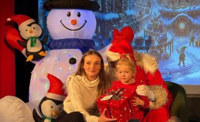 Na zdjęciu widać dziewczynkę z prezentem w dłoniach i kobietę. Za nimi znajduje się mężczyzna przebrany za Mikołaja. Wokół panuje świąteczna sceneria. 