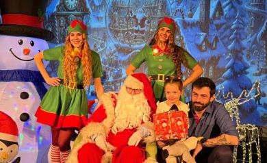 Dziecko i mężczyzna pozujący do zdjęcia z mężczyzną przebranym za Mikołaja i dwoma kobietami przebranymi za elfy. W tle widać świąteczną scenerię. 
