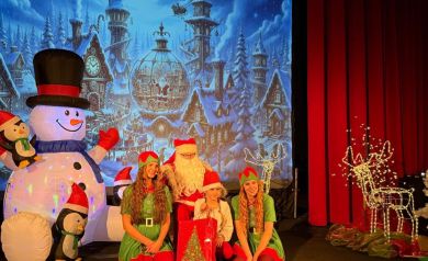 Dziecko pozujące do zdjęcia z mężczyzną przebranym za Mikołaja i dwoma kobietami przebranymi za elfy. W tle widać świąteczną scenerię. 