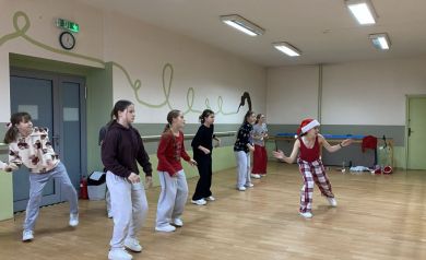 Zdjęcie przedstawia grupę młodych ludzi, dziewcząt, uczestniczących w zajęciach tanecznych w sali tanecznej. Jedna z kobiet ma na głowie świąteczną czapkę.