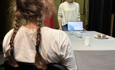 Na zdjęciu dwie dziewczyny. Jedna z nich siedzi tyłem. Między nimi znajduje się stół nakryty białym obrusem. Stoją na nim filiżanki, kubki, talerzyk z ciastkami oraz laptop. 