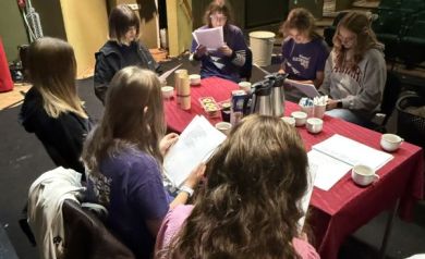 Młodzież siedząca na scenie przy stole, który jest nakryty czerwonym obrusem. Wpatrują się w kartki trzymane w rękach. 