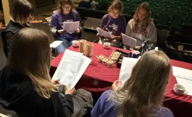 Młodzież siedząca na scenie przy stole, który jest nakryty czerwonym obrusem. Na stole znajdują się ciasteczka, filiżanki, termosy, karton mleka i białe kartki papieru. Wpatrują się w kartki trzymane w rękach. 