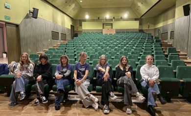 Siedem dziewcząt pozujących do zdjęcia. Siedzą na widowni. Za nimi bardzo duża ilość zielonych krzesełek.