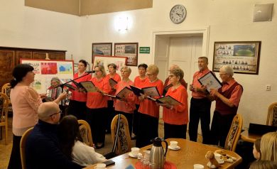 Chór osób starszych występuje w sali spotkań, prowadzony przez dyrygentkę, większość chórzystów ubrana jest w czerwone lub koralowe bluzki i ciemne spodnie lub spódnice, natomiast mężczyźni mają koszule i kamizelki w podobnych kolorach. Przed nimi siedzi publiczność przy stołach nakrytych do poczęstunku.