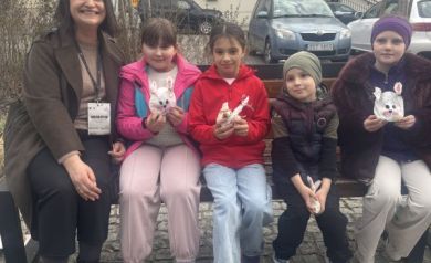 Uśmiechnięta czwórka dzieci trzyma znalezione torebki niespodzianki w kształcie zajączka, jest z nimi uśmiechnięta kobieta w brązowym płaszczu. Wszyscy siedzą na ławce.